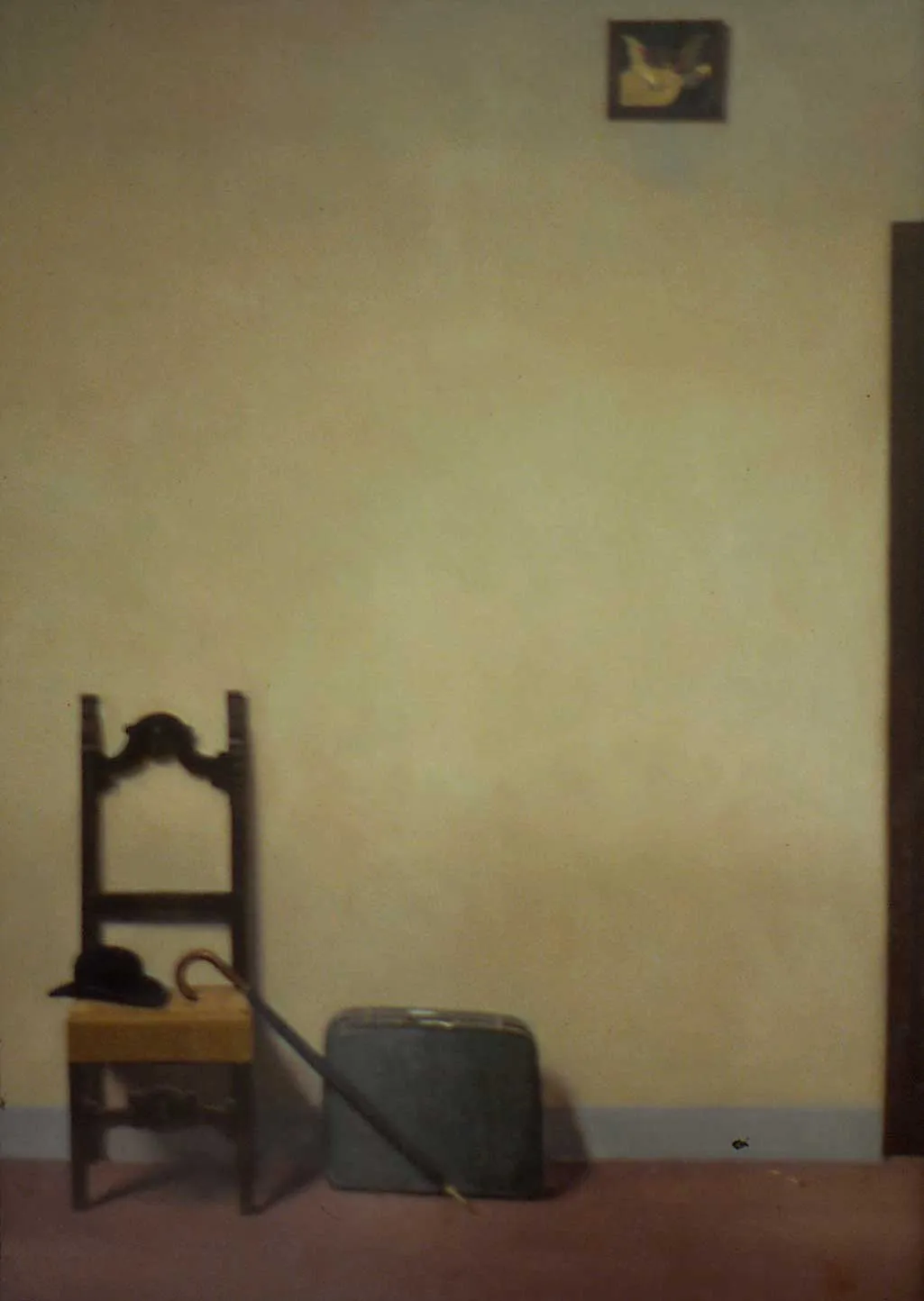 High on a wall a painting of putti looks down on a chair, suitcase and umbrella. 